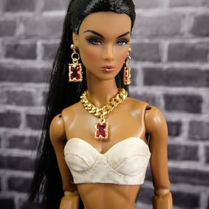 May include: A doll wearing gold-toned jewelry with red accents, including a necklace and earrings. The doll has dark hair and is wearing a white bustier and dark pants. The background is a gray brick wall.