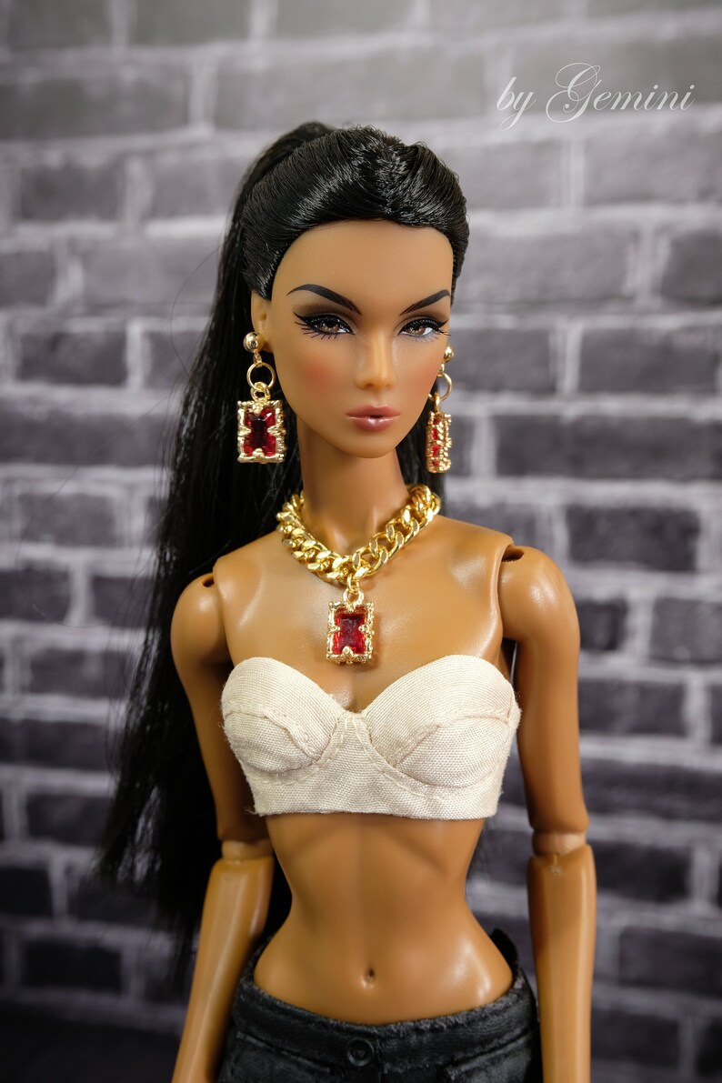 May include: A doll wearing gold-toned jewelry with red accents, including a necklace and earrings. The doll has dark hair and is wearing a white bustier and dark pants. The background is a gray brick wall.