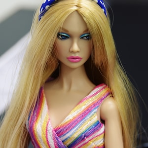 May include: A doll with long blonde hair wearing a blue and white polka dot headband and a colorful striped dress.