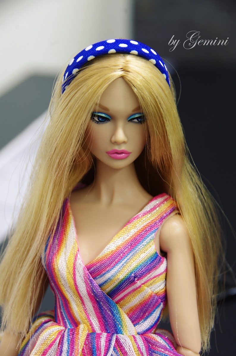 May include: A doll with long blonde hair wearing a blue and white polka dot headband and a colorful striped dress.