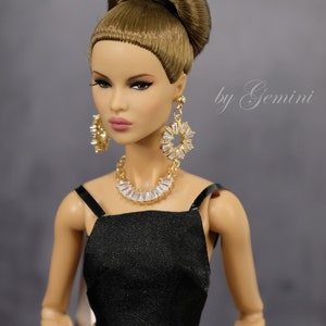 Pu&ograve; includere: Una bambola di moda con i capelli castani raccolti in un alto chignon, indossa un top nero senza maniche, pantaloni kaki con cintura, orecchini dorati e una collana di cristalli. La bambola ha un'espressione neutra e guarda la telecamera. Il testo "by Gemini" &egrave; scritto in corsivo sullo sfondo.