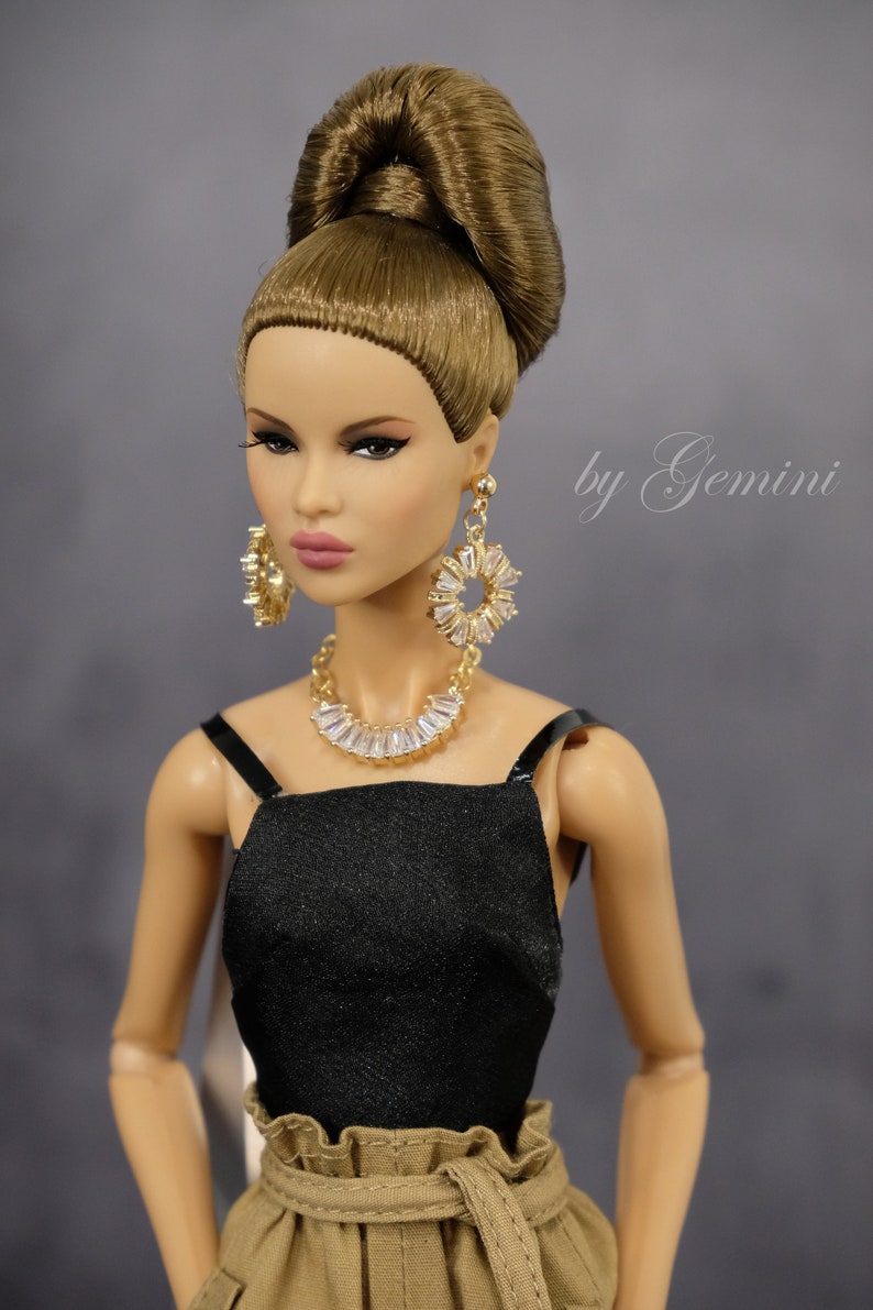 Pu&ograve; includere: Una bambola di moda con i capelli castani raccolti in un alto chignon, indossa un top nero senza maniche, pantaloni kaki con cintura, orecchini dorati e una collana di cristalli. La bambola ha un'espressione neutra e guarda la telecamera. Il testo "by Gemini" &egrave; scritto in corsivo sullo sfondo.