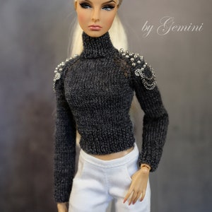 May include: A doll wearing a gray knitted turtleneck sweater with silver embellishments on the shoulders and white pants. The doll has blonde hair and is wearing makeup.