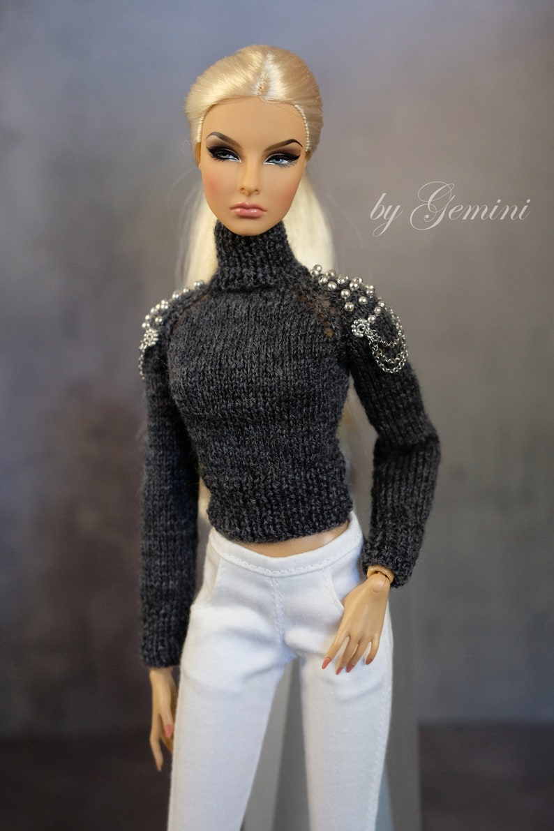 May include: A doll wearing a gray knitted turtleneck sweater with silver embellishments on the shoulders and white pants. The doll has blonde hair and is wearing makeup.