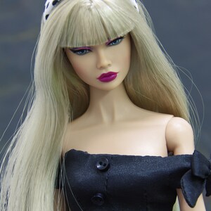 May include: A doll with blonde hair and a black and white polka dot headband is wearing a black off-the-shoulder dress with black buttons. The doll has pink lips and green eyes.