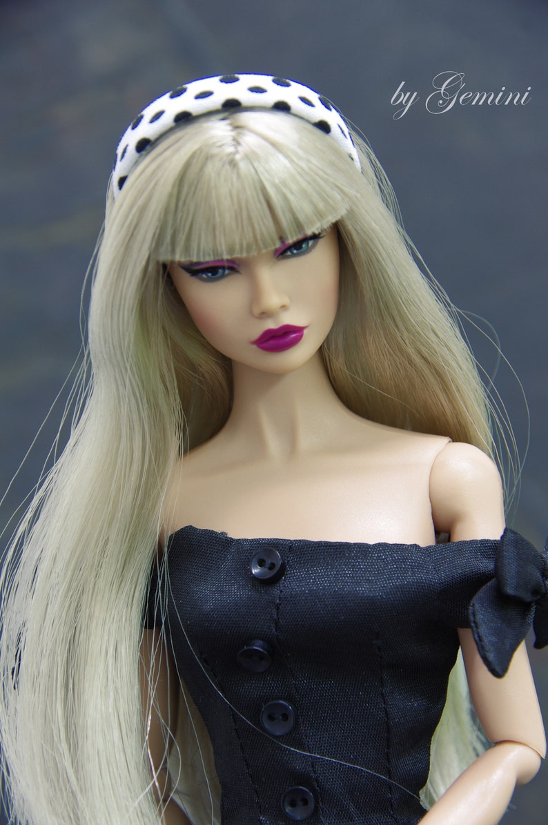 May include: A doll with blonde hair and a black and white polka dot headband is wearing a black off-the-shoulder dress with black buttons. The doll has pink lips and green eyes.