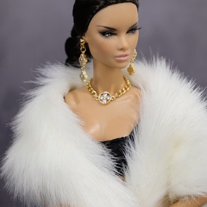 Pu&ograve; includere: Una bambola che indossa un cappotto di pelliccia bianca, una collana d'oro con un grande ciondolo di pietra chiara e orecchini d'oro. La bambola ha i capelli scuri e il trucco.