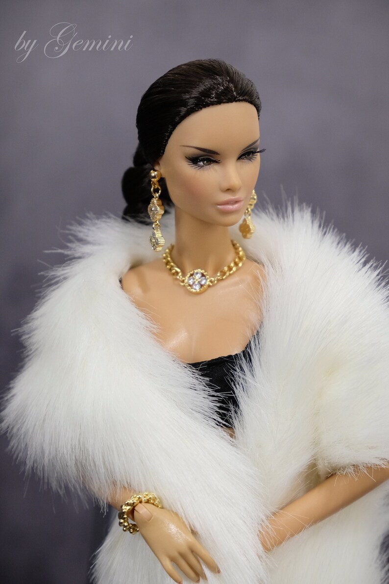 Pu&ograve; includere: Una bambola che indossa un cappotto di pelliccia bianca, una collana d'oro con un grande ciondolo di pietra chiara e orecchini d'oro. La bambola ha i capelli scuri e il trucco.