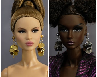 May include: Two fashion dolls with different hairstyles and makeup. The doll on the left has long brown hair in a bun and is wearing gold earrings. The doll on the right has short black hair with two buns and is wearing silver earrings.