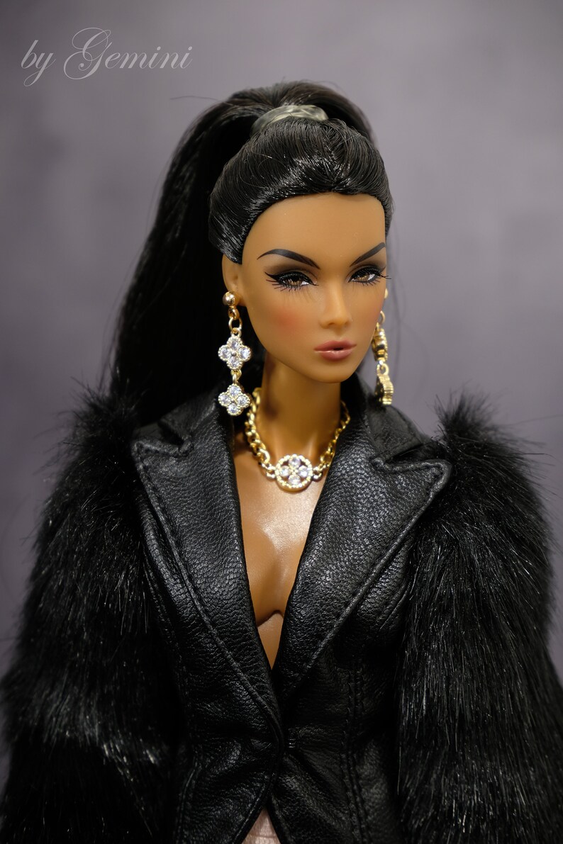 Pu&ograve; includere: Una bambola di moda con lunghi capelli neri raccolti in una coda di cavallo, indossa una giacca di pelle nera con bordi in pelliccia, una collana d'oro con un ciondolo a forma di diamante e orecchini di diamanti.