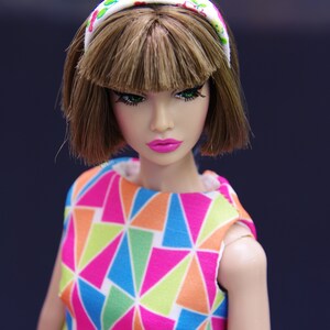 May include: A doll with brown hair and green eyes wearing a white headband with pink and green flowers and a colorful geometric print dress.