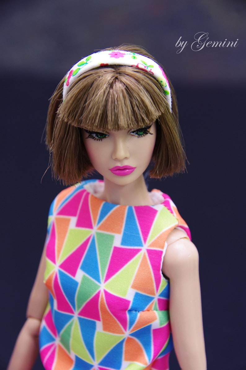 May include: A doll with brown hair and green eyes wearing a white headband with pink and green flowers and a colorful geometric print dress.