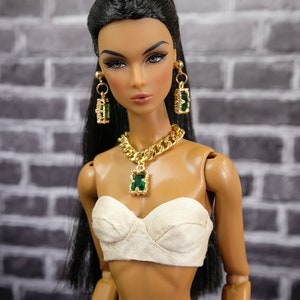 May include: A doll wearing a gold chain necklace with a green pendant, matching earrings, and a white strapless top. The doll has long black hair and is wearing dark denim pants. The background is a gray brick wall.