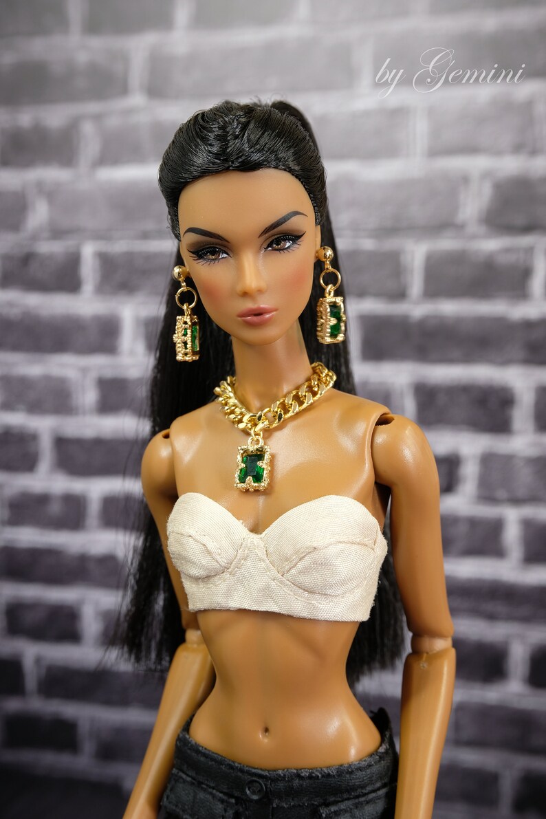 May include: A doll wearing a gold chain necklace with a green pendant, matching earrings, and a white strapless top. The doll has long black hair and is wearing dark denim pants. The background is a gray brick wall.