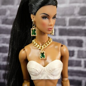 May include: A fashion doll with long black hair in a high ponytail, wearing a gold necklace, earrings, and a matching pendant with green rectangular stones. The doll is also wearing a white bustier and dark denim pants.