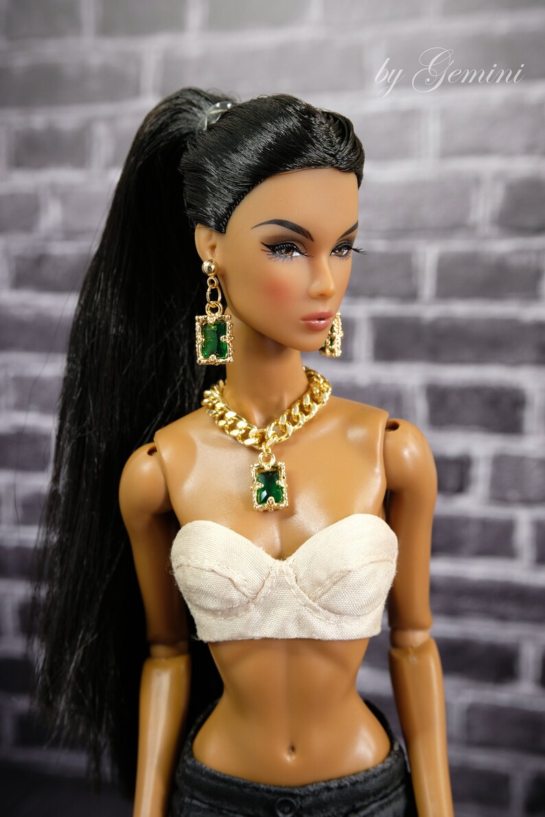 May include: A fashion doll with long black hair in a high ponytail, wearing a gold necklace, earrings, and a matching pendant with green rectangular stones. The doll is also wearing a white bustier and dark denim pants.