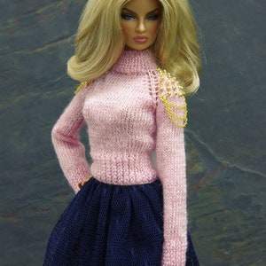 May include: A doll wearing a pink knitted sweater with gold chain detailing on the shoulder and a navy blue tulle skirt. The doll has long blonde hair and blue eyes.