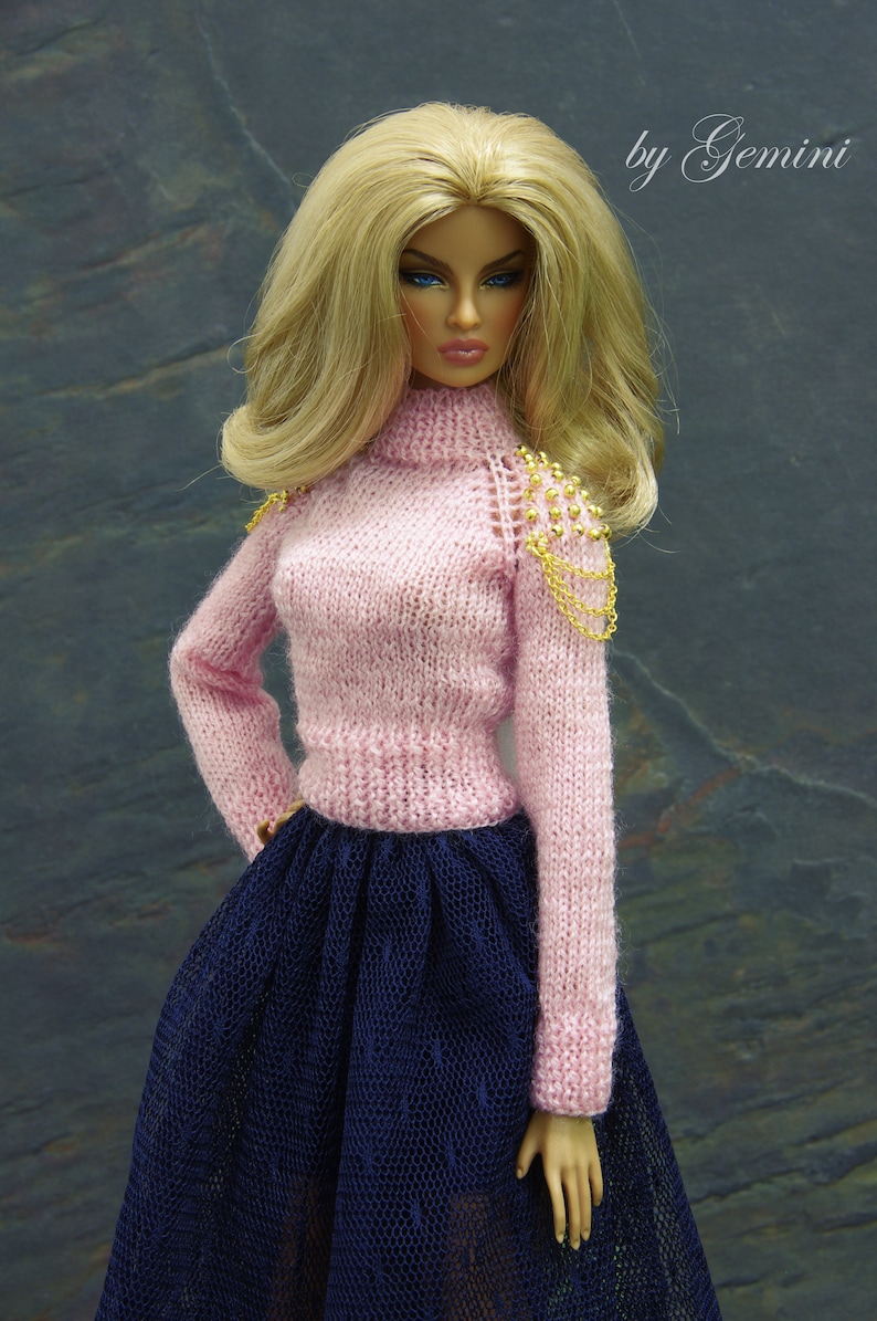 May include: A doll wearing a pink knitted sweater with gold chain detailing on the shoulder and a navy blue tulle skirt. The doll has long blonde hair and blue eyes.