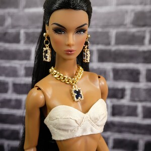 May include: A fashion doll with dark hair and makeup, wearing a cream-colored top, and a gold chain necklace with a blue pendant. The doll also has matching gold earrings. The background is a gray brick wall. The text "by Gemini" is in the upper right corner.