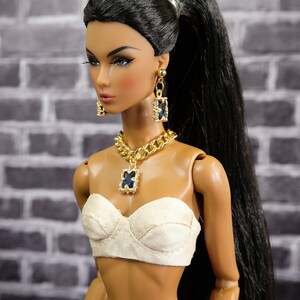 May include: A fashion doll with long black hair in a high ponytail, wearing a cream-colored bustier, and dark denim pants. The doll is accessorized with a gold chain necklace and matching earrings, each featuring a dark blue square pendant. The image is signed "by Gemini".