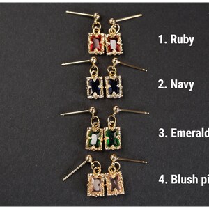 May include: Gold-toned stud earrings with rectangular gemstones in ruby, navy, emerald, and blush pink. Each pair features a small, dangling rectangular stone surrounded by smaller accent stones. The image is on a black background.