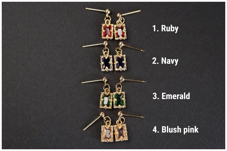 May include: Gold-toned stud earrings with rectangular gemstones in ruby, navy, emerald, and blush pink. Each pair features a small, dangling rectangular stone surrounded by smaller accent stones. The image is on a black background.