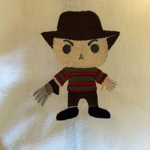 May include: Embroidered image of a cartoon character with a brown hat, a striped sweater, and a glove with sharp claws. The character is depicted with a neutral expression and black shoes. The embroidery is on a cream-colored fabric.