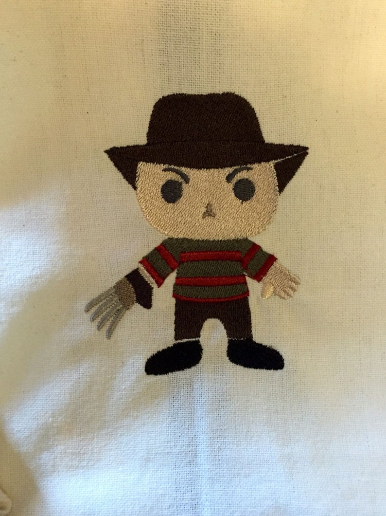May include: Embroidered image of a cartoon character with a brown hat, a striped sweater, and a glove with sharp claws. The character is depicted with a neutral expression and black shoes. The embroidery is on a cream-colored fabric.