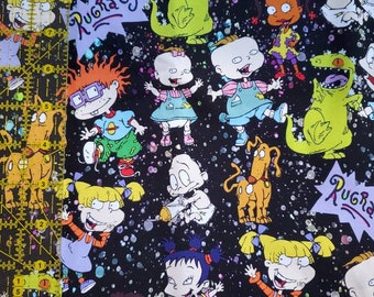 May include: Fabric featuring a repeating pattern of cartoon characters from the Rugrats animated series. The design includes characters like Tommy, Chuckie, Angelica, and Reptar, set against a black background with white speckles. The word "Rugrats" is also visible.