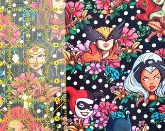 May include: A black fabric with white polka dots and illustrations of female comic book characters, surrounded by pink and orange flowers. A clear Omnigrid ruler with yellow markings is on the left side of the image.