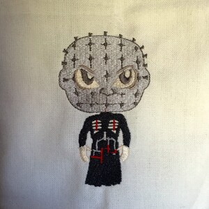 May include: Embroidered design of a character with a gray head covered in pins, large brown eyes, and a black outfit with red accents. The character has white gloves and is set against a light background.