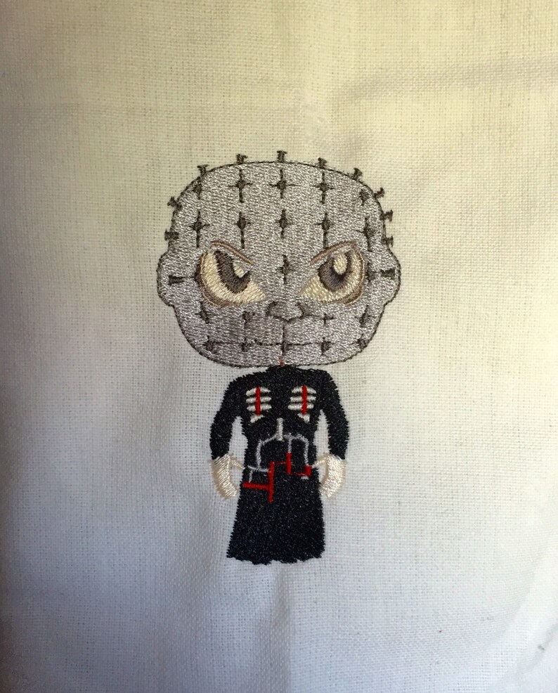 May include: Embroidered design of a character with a gray head covered in pins, large brown eyes, and a black outfit with red accents. The character has white gloves and is set against a light background.