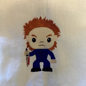 May include: An embroidered figure of a character from a horror movie, with orange hair, a blue jumpsuit, and a bloody knife. The figure has a stern expression and is set against a white background.