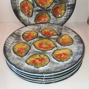 May include: A set of six white ceramic oyster plates with black and gray striped edges. Each plate has six indented sections with a yellow and red design.