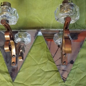 Puede incluir: Un par de apliques de pared con placas traseras de espejo triangulares y brazos de metal curvados en tono bronce. Cada lámpara está rematada con un soporte de cristal transparente y facetado, ofreciendo un diseño reflectante y estructurado.