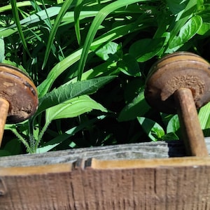 May include: Two wooden spools with brown paint and a worn finish, attached to a wooden board. The spools are mounted on metal rods.