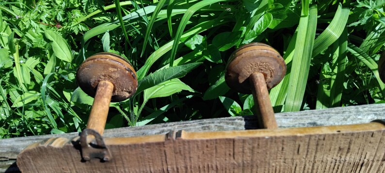 May include: Two wooden spools with brown paint and a worn finish, attached to a wooden board. The spools are mounted on metal rods.