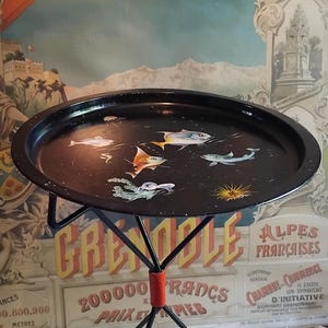 Könnte beinhalten: Ein runder schwarzer Metall-Tabletttisch mit einer bemalten Unterwasserwelt-Szene, die verschiedene Fische, eine Qualle und einen Seeigel zeigt. Das Tablett ruht auf drei dünnen schwarzen Metallbeinen, die durch ein orangefarbenes Verbindungsstück verbunden sind.