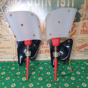 Puede incluir: Un par de apliques de pared vintage con detalles en negro y rojo. Las apliques tienen pantallas blancas con detalles dorados y están montadas sobre bases de metal negro.