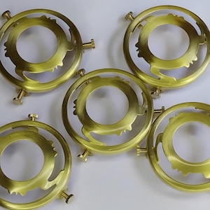 Può includere: Cinque anelli circolari per paralumi color ottone, con intagli decorativi e fissaggi a vite. Gli anelli hanno un'apertura centrale e sono disposti su una superficie bianca. Il design presenta un volto stilizzato.