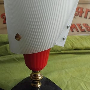 May include: A table lamp featuring a white, ribbed, conical shade adorned with small gold-toned pyramid studs. The lamp has a bright red, fluted base section, a polished gold-toned neck, and a black, three-legged, tripod-style base stand.