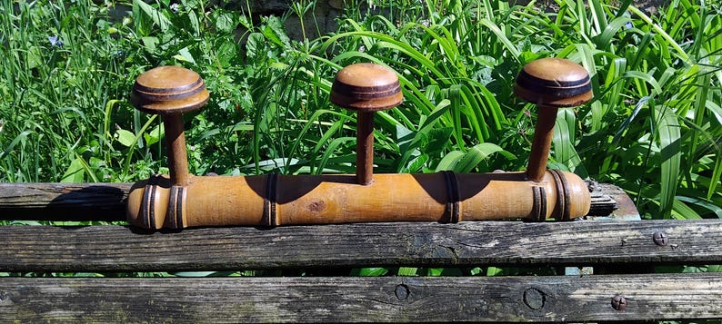 May include: A wooden coat rack with three hooks. The rack is made of a single piece of wood with a natural brown finish. The hooks are round and have a dark brown finish.