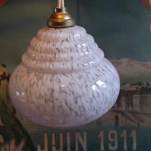 Puede incluir: Una lámpara colgante vintage con una pantalla de vidrio blanco y gris veteado. La pantalla tiene un diseño único en capas y está suspendida de un accesorio de color latón. El fondo presenta un póster vintage con el texto "JUIN 1911".