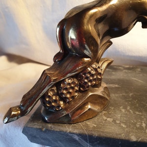 May include: A gold-toned metal sculpture of a deer with its head turned back, standing on a black marble base. The deer is depicted with its legs bent and its tail raised, as if it is leaping. The sculpture is detailed with textured fur and a delicate, curved horn.