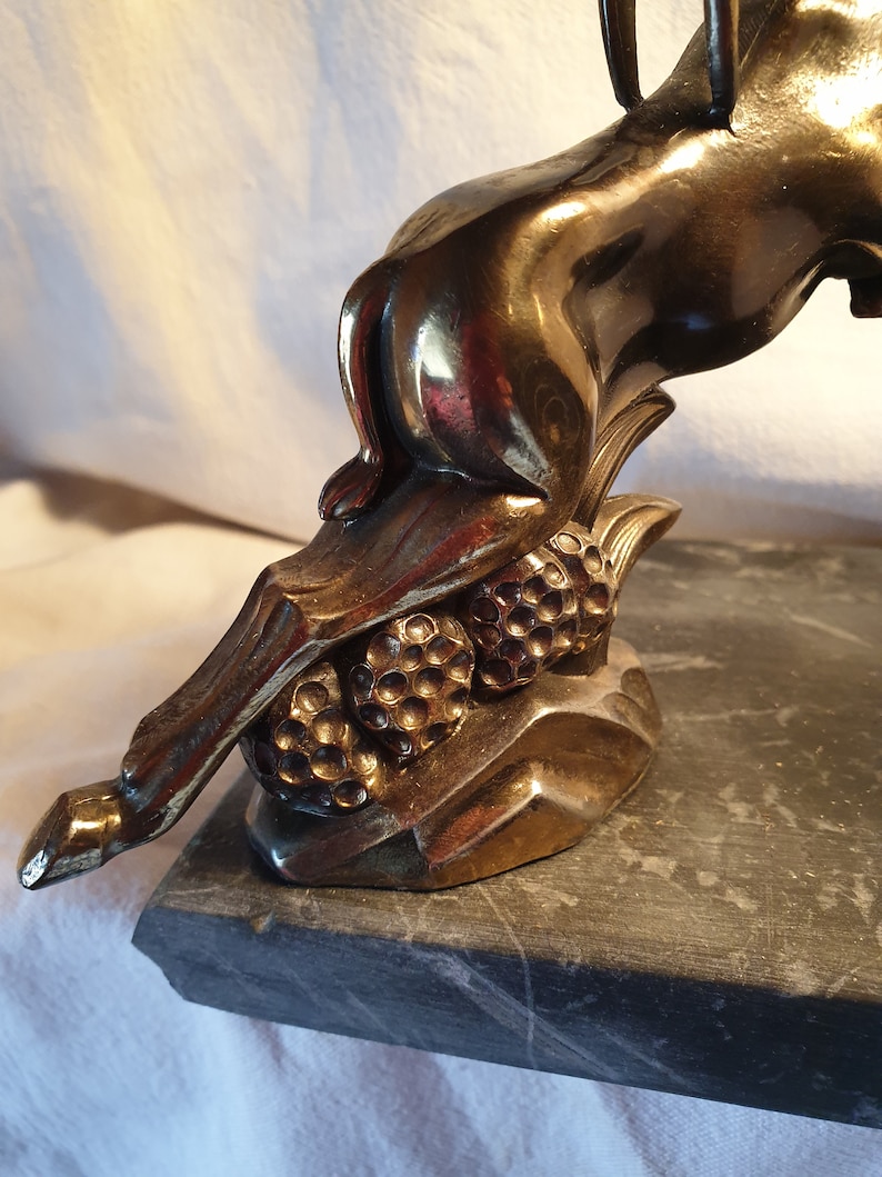 May include: A gold-toned metal sculpture of a deer with its head turned back, standing on a black marble base. The deer is depicted with its legs bent and its tail raised, as if it is leaping. The sculpture is detailed with textured fur and a delicate, curved horn.