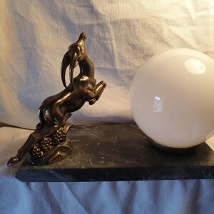 May include: A vintage table lamp with a brass gazelle sculpture and a white glass globe shade. The lamp sits on a black marble base.