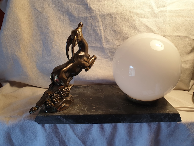 May include: A vintage table lamp with a brass gazelle sculpture and a white glass globe shade. The lamp sits on a black marble base.