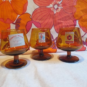 May include: Three amber glass stemmed drinking glasses with labels. The glasses are on a white surface with a floral pattern in the background. The labels read "Cordial Medoc", "Beefeater London Distilled Dry Gin", and "Dubonnet".