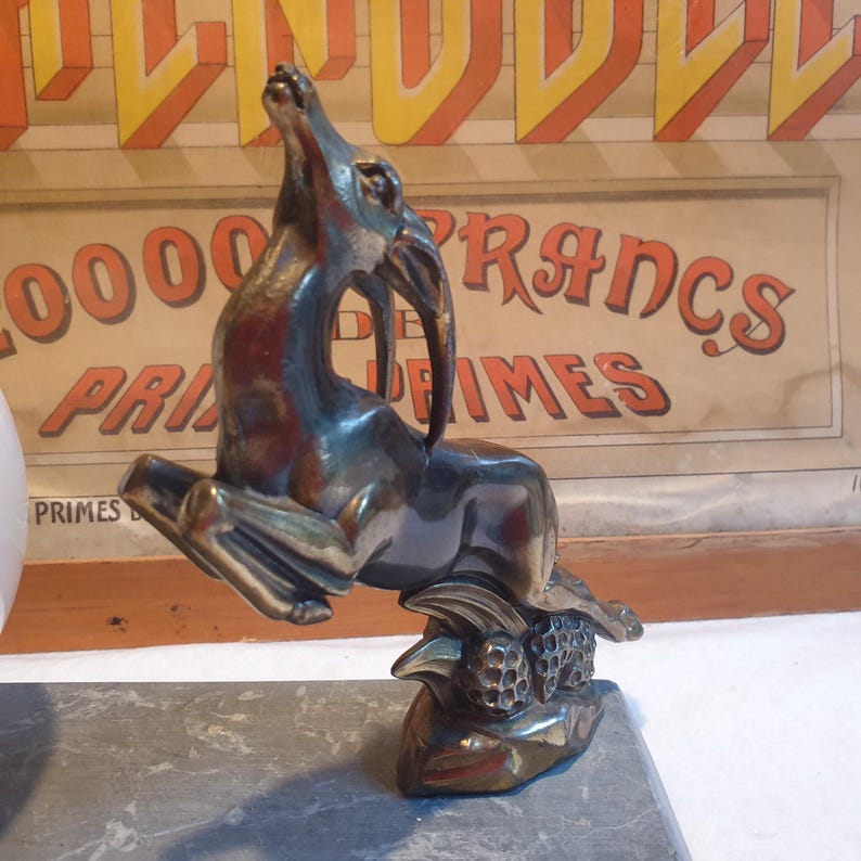 May include: A bronze sculpture of a leaping antelope with its head tilted back. The antelope is standing on a base with a textured surface. The sculpture is on a gray stone surface.