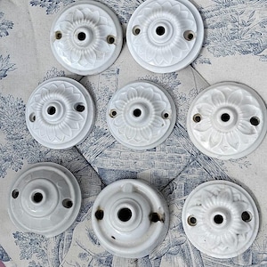 Pode incluir: Uma coleção de oito placas elétricas circulares de cerâmica branca, cada uma com um orifício central e dois orifícios laterais menores para montagem. As placas apresentam um padrão floral em relevo semelhante a pétalas.